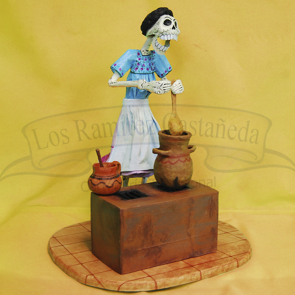 Personaje en calavera de una mujer cocinando en un fogón de tierra. Sobre el fogón hay dos ollas de barro.